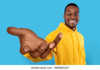 Happy African American guy showing big outstretched hand, offering help, taking or giving something, reaching out for support on blue studio background. Selective focus
