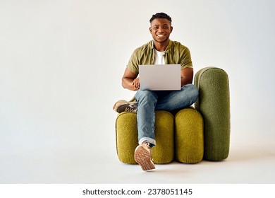 Happy african american guy sitting on design chair and holding wireless laptop. Male freelancer in casual attire looking at camera with toothy smile over white background. - Powered by Shutterstock