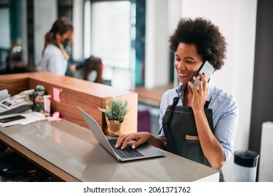 Happy African American Hairdresser Working On Laptop While Talking On Mobile Phone At Hair Salon. 