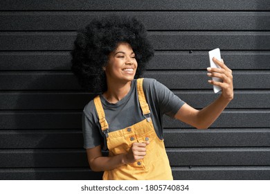 Happy African American hipster woman blogger with Afro hair laughing, holding phone, recording vlog, shooting social media stories or video calling in mobile app standing on black background.