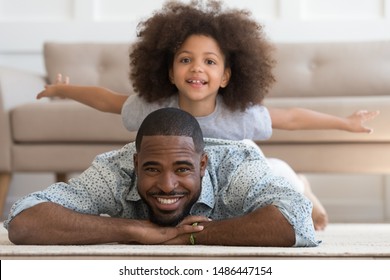 Happy African American Joyful Family Of Two Portrait. Cheerful Smiling Black Father Lying On Floor Carpet, Carrying Little Cute Mixed Race Daughter, Pretending To Be Plane, On Back Looking At Camera.