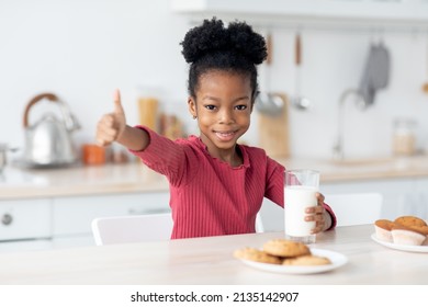 Happy African American Kid Drinking Milk, Showing Thumb Up. Cute Little Black Girl Sitting At Table, Having Snack, Holding Glass Of Milk, Eating Cookies, Kitchen Interior, Copy Space