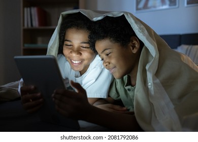 Happy African American kids lying under duvet and reading online book on tablet in dark room - Powered by Shutterstock