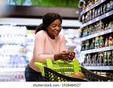 Happy African American lady buying food, checking grocery list on smartphone at supermarket. Positive black woman with mobile device and trolley purchasing products at mall