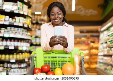 Happy African American Lady Doing Grocery Shopping On Mobile Phone Via Application, Posing With Cellphone And Cart In Supermarket. Woman Advertising Shop Buying Food And Using Smartphone