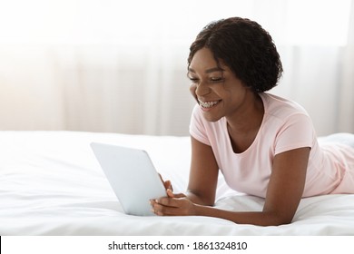 Happy african american lady enjoying weekend, laying with pad on bed, using social media or websurfing. Smiling young black woman using digital tablet while resting at home, empty space. - Shutterstock ID 1861324810