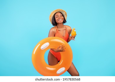 Happy African American lady with inflatable ring and tasty tropical cocktail smiling at camera on blue studio background. Cheerful black woman having fun during her summer beach vacation