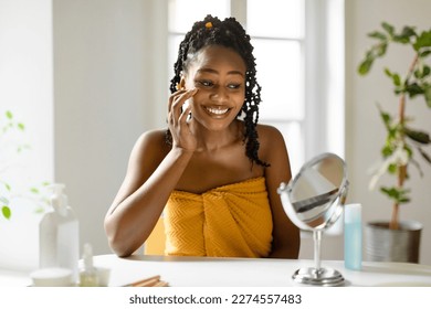 Happy african american lady looking at mirror and applying under eye cream, sitting wrapped in towel, making beauty treatments at home, enjoying self-care, free space - Powered by Shutterstock