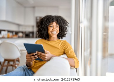 Happy African American lady using tab device looking away on sofa at home. Smiling pretty young woman sitting on couch relaxing looking away at window holding digital table in kitchen.