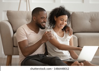 Happy african american loving family couple hugging, sitting on warm heated floor in living room, holding computer, watching photos from vacation or honeymoon, enjoying funny videos together.