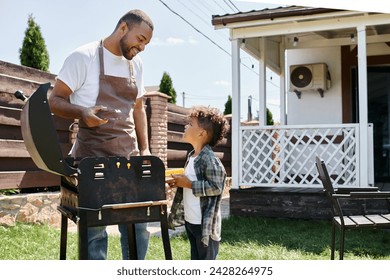 happy african american man in apron cooking corn on bbq grill and looking at son on backyard - Powered by Shutterstock