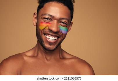Happy African American man with bare shoulders and LGBT flag painted on cheeks smiling and looking at camera against brown background - Shutterstock ID 2211068575