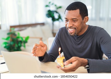 Happy African American Man Celebrating Birthday At Home During Pandemic Quarantine Time.He Blows Cake With Friends On Through Video Call Virtual Party With Laptop Computer. Coronavirus Outbreak.