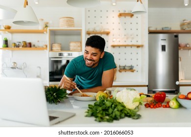 Happy African American Man Cooking Healthy Dinner At Home. He Is Following A Video Tutorial On The Laptop.