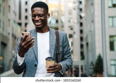 Happy african american man in earphones using mobile phone while drinking coffee on city street