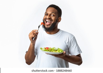 Happy African American Man Eating Healthy Salad For Dinner Standing Posing Over White Background In Studio. Male Weight Loss, Dieting Nutrition Concept. Black Guy Enjoying Veggie Meal