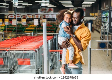 happy african american man embracing and holding little daughter in grocery store