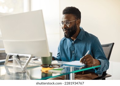 Happy african american man entrepreneur sitting at worktable, looking at computer screen and taking notes at office, looking for new business opportunities, copy space - Powered by Shutterstock