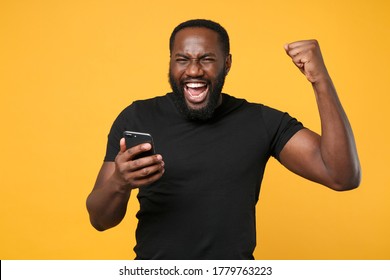 Happy African American Man Football Fan In Casual Black T-shirt Isolated On Yellow Background Studio Portrait. People Lifestyle Concept. Mock Up Copy Space. Using Mobile Phone Doing Winner Gesture