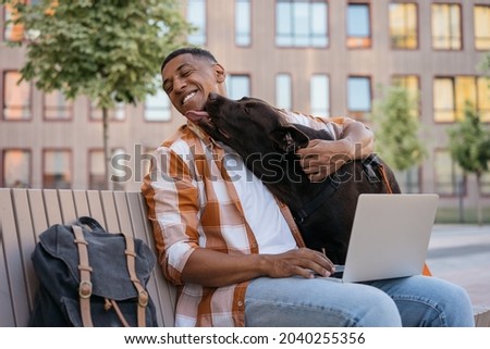 Happy African American man freelancer using laptop computer, working online, hugging with adorable pet. Labrador dog licking his owner in park, selective focus. Best friends concept 