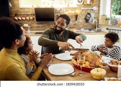 Happy African American Man Having Thanksgiving Lunch With His Family And Carving Stuffed Turkey At Dining Table.