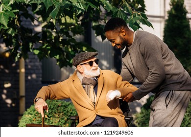 Happy African American Man Helping Senior Disabled Man To Get Up From Wheelchair On Street