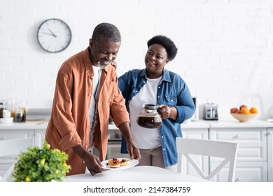 happy african american man holding pancakes on plate near wife with coffee pot - Powered by Shutterstock