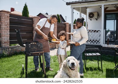 happy african american man serving grilled corn on plate of son near wife and dog on backyard - Powered by Shutterstock