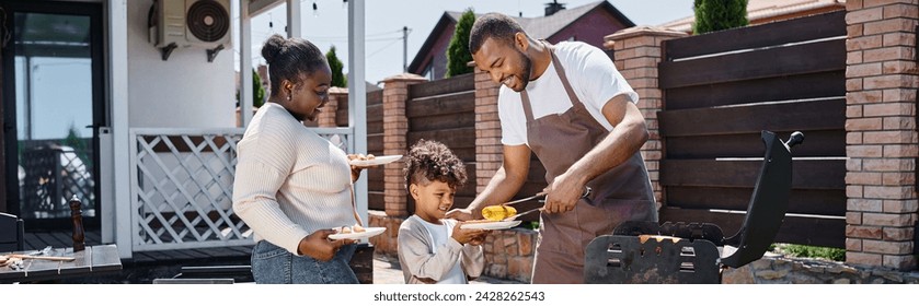 happy african american man serving grilled corn on plate of son near wife during bbq party, banner - Powered by Shutterstock