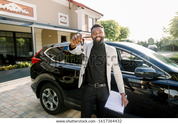 happy African American man\
showing car key standing near new black car crossover outdoors.\
Young African American businessman holding a car key and car sales\
contract