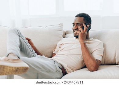 Happy African American man sitting on a black sofa, using his smartphone for a video call while relaxing at home He is confidently chatting and smiling, enjoying the modern technology and wireless - Powered by Shutterstock