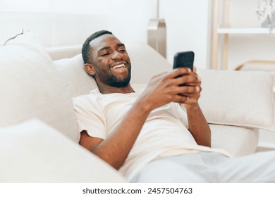 Happy African American man sitting on a black sofa in a modern apartment, holding a mobile phone and typing a message He is relaxed and enjoying his leisure time, using wireless technology to connect - Powered by Shutterstock