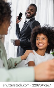 Happy African American Man In Suit Holding Smartphone And Cup Of Coffee Near Family On Blurred Foreground