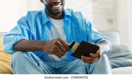 Happy African American Man Taking Credit Card Out Of Wallet, Sitting On Couch In Living Room Interior, Selective Focus, Closeup, Panorama