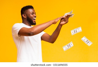 Happy african american man throwing out money banknotes, yellow studio background