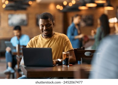 Happy African American man using laptop while sitting in a bar.  - Powered by Shutterstock