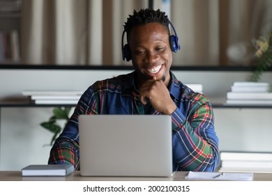 Happy African American man in wireless headphones looking at laptop screen, excited young student watching webinar, engaged in online educational course, intern studying in office, sitting at desk