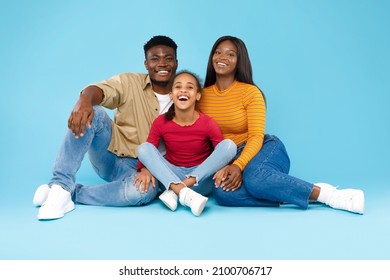 Happy African American man, woman and girl sitting on the floor isolated on blue studio wall. Smiling laughing daughter posing with mom and dad looking at camera. Healthy Family Relationship Concept