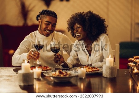 Happy african american married couple having dinner by candlelight at home and toasting with wine glasses. Black romantic couple is drinking wine and smiling. Celebrating their love at home.