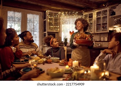 Happy African American mature woman brining stuffed turkey at dining table during family Thanksgiving dinner.