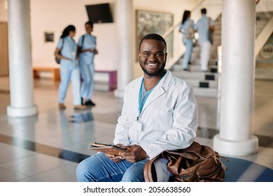 Happy African American Medical Student In White Lab Coat Sitting In Corridor At Medical University And Looking At Camera.