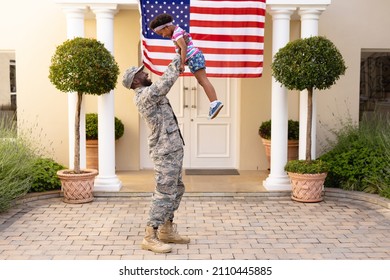 Happy african american military father holding aloft daughter in front of house with usa flag. family, bonding and patriotism, unaltered.