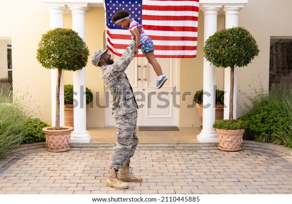 Happy african american military father holding\
aloft daughter in front of house with usa flag. family, bonding and\
patriotism, unaltered.