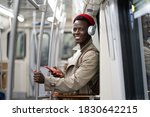 Happy African American millennial man in red hat, trench coat stand in subway train, using mobile phone, listens to music with wireless headphones in public transportation.  