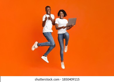 Happy African American Millennial Man And Woman Jumping Up With Laptop, Shouting And Gesturing On Orange Studio Background. Young Black Couple Gambling Online Successfully, Using Laptop, Free Space