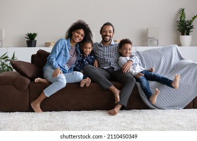 Happy African American Millennial Parents And Sweet Little Preschool Sibling Children Resting On Home Sofa, Hugging, Looking At Camera, Smiling. Black Family With Two Kids Full Length Portrait