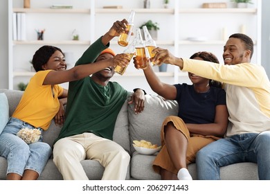 Happy African American Millennials Two Beautiful Couple Enjoying Home Party, Cheerful Black Men And Women Sitting On Couch In Living Room, Drinking Beer, Eating Snacks, Celebrating Weekend