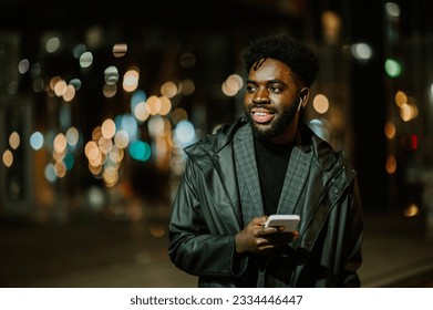 A happy African American modern man is using his phone for a call on the city street downtown at night. A man is using his phone on the street at night while looking away. - Shutterstock ID 2334446447