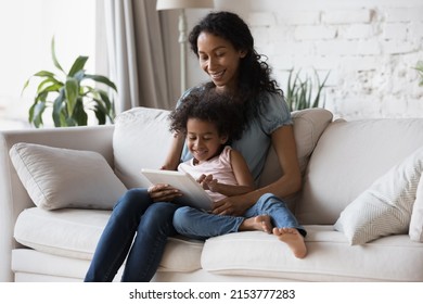 Happy African American mom holding sweet daughter on lap, teaching kid to use learning application on tablet. Mother and kid enjoying leisure at home, reading ebook on digital pad