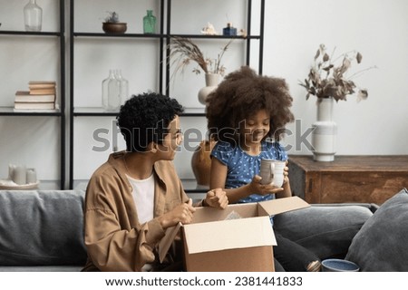 Happy African American mom and pre teen daughter girl unpacking moving box, relocating into new apartment, producing dish. Customers opening parcel ceramic mugs, producing cups from carton container